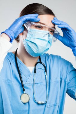 Genç harap olmuş kadın NHS Genel Uygulayıcı doktor yüz maskesi ve koruyucu gözlük takıyor, elleri başında, Coronavirus 'un artması, COVID-19 salgını yüzünden birçok hastanın ölümü,