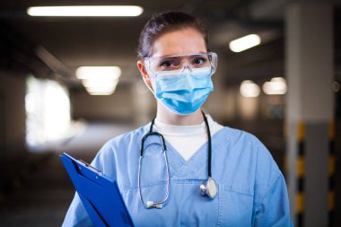 Genç bayan NHS doktoru klinikte, hastane koridorunda ya da koridorda, acil hasta otoparkında giriş alanında, koruyucu maske takan ve gözlük takan personel portresi