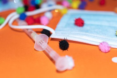 Vaduz, Lihtenştayn, 17 Aralık 2021 Turuncu arka planda virüs parçacıkları ve şırıngalarla tıbbi yüz maskeleri