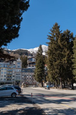 Davos, İsviçre, 23 Mart 2022 Güneşli bir günde arka planda ağaçların ve binaların bulunduğu bir sokak ve küçük bir parka bakın.
