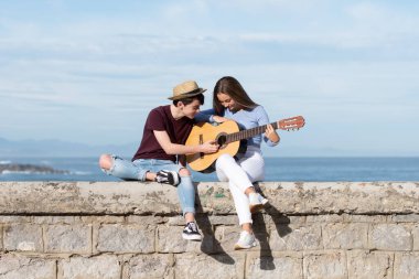 Genç çocuk kız arkadaşına deniz kenarında gitar çalmayı öğretiyor.