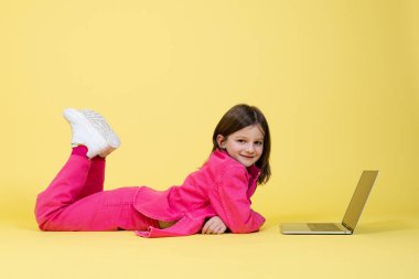 Tatlı küçük kız dizüstü bilgisayarın önünde yerde yatıyor ve kameraya bakıyor.