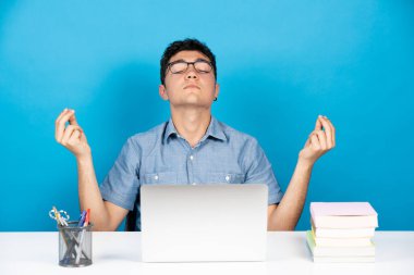 İspanyol üniversite öğrencisi dizüstü bilgisayarın önünde meditasyon yaparken mavi arka planda izole edilmiş.