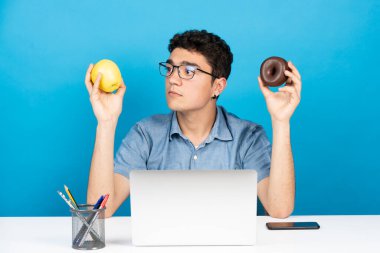 İspanyol adam elmaya bakıyor ve elinde çikolatalı donut tutuyor. Diğer yandan mavi arka planda izole edilmiş.