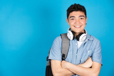 İspanyol üniversite öğrencisi mavi arka planda izole edilmiş kameraya bakıyor. Kollarını kavuşturmuş bir gencin portresi.