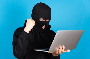 Siyah kar maskesi takan hacker, mavi arkaplanda dizüstü bilgisayarını tutarken yeni siber suçu kutluyor..