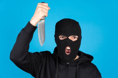 Angry assasin wearing black ski mask and holding and menacing with knife isolated on blue background. Delinquent looking at camera