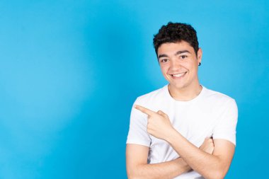 Esmer Latin kökenli genç çocuk, yan fotokopi alanını gösteriyor ve mavi arka planda izole edilmiş kameraya bakıyor. Genç adam gülümsüyor