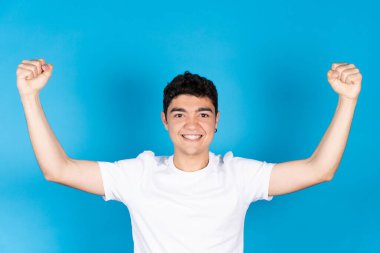 Mavi arka planda kollarını yukarda gösteren mutlu İspanyol genç çocuk. Sağlıklı yaşam tarzı kavramı