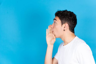 Latin kökenli genç bir çocuğun mavi arka planda izole edilmiş alanı kopyalamak için gizli bir eli ağıza verdiği yan görüş..