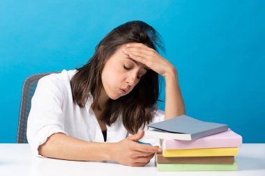 Hispanic young woman despaired in front of pile of books isolated on blue background. Student with stress before exam.