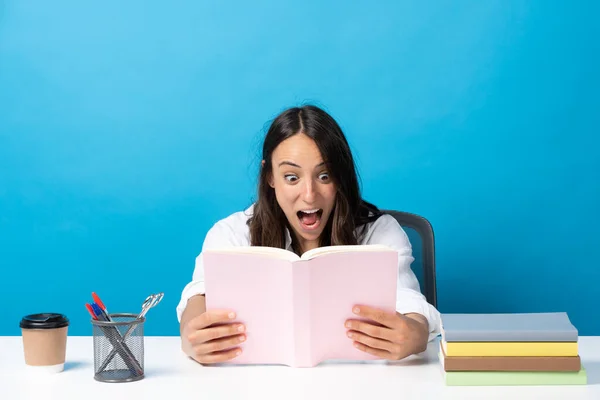 Surprised Hispanic Woman Looking Book Sitting Desk Isolated Blue Background — Zdjęcie stockowe