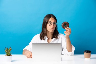 Laptopun önünde çikolatalı çöreğe bakan İspanyol genç kadın mavi arka planda izole edilmiş. İş konseptinde kötü beslenme alışkanlıkları