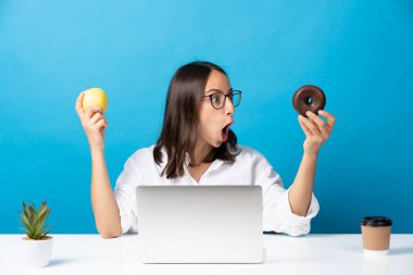 Şaşırmış İspanyol kadın çikolatalı çöreğe bakıyor ve diğer elinde mavi arka planda izole edilmiş bir elma tutuyor..