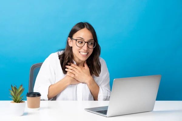 Mavi arka planda izole edilmiş görüntülü konuşma sırasında İspanyol genç kadın ellerini kalbe koydu. İş kadını dizüstü bilgisayarın önünde
