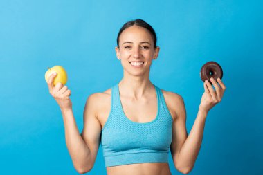 Ellerinde elmalı ve çikolatalı çörek tutan İspanyol sporcu kadın mavi arka planda izole edilmiş kameraya bakıyor..