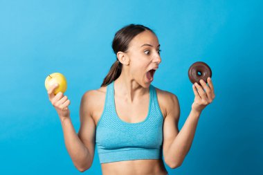 Süpriz sporcu kadın çikolatalı çöreğe bakıyor ve mavi arka planda elma tutuyor. Tatlı yemek cazibesi ve bağımlılık konsepti