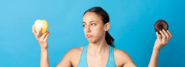 Üzgün İspanyol zinde kadın elmaya bakıyor ve mavi arka planda çikolatalı çörek tutuyor..