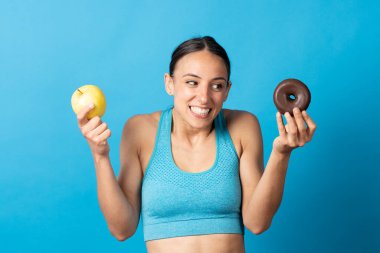 İspanyol sporcu kadın çikolatalı çöreğe bakarken mavi arka planda izole edilmiş meyveleri tutuyor..