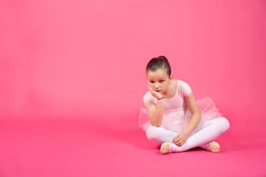 Sıkıcı küçük balerin kız yerde oturmuş kameraya bakıyor. Canlı pembe arkaplanlı stüdyo görüntüsü.