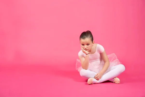 床に座ってカメラを見ている小さなバレエダンサーの女の子を退屈 スタジオショットともに鮮やかなピンクの背景 — ストック写真
