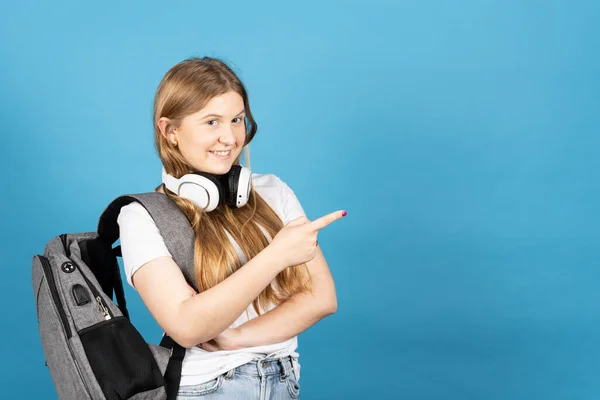 Pretty female student pointing at copy space isolated on blue background. Teenager girl wearing headphones and backpack.