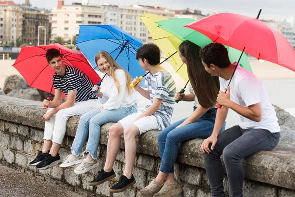 Grupa Nastolatków Rozmawia Trzymając Kolorowe Parasole Przyjaciele Siedzący Ścianie — Zdjęcie stockowe