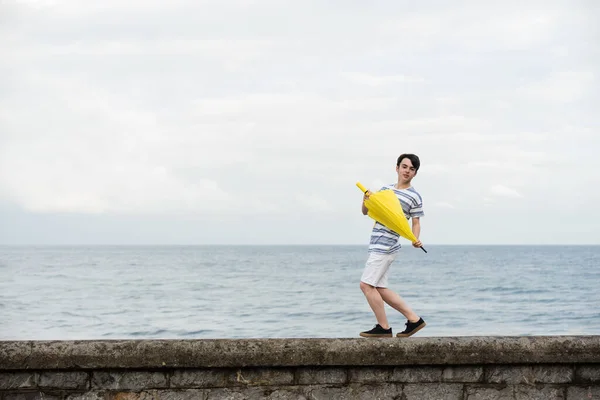 在雨中 一个可爱的少年拿着一把黄色的雨伞 在海滨的墙上跳舞 — 图库照片