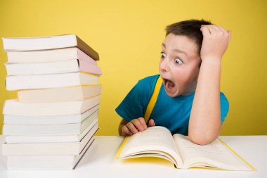 Scared student boy looking at pile of books. Overwhelmed schoolboy isolated on yellow background.
