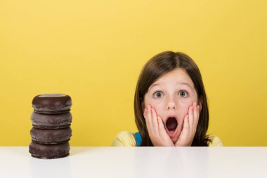 Surprised little girl beside pile of chocolate doughnut isolated on yellow background.