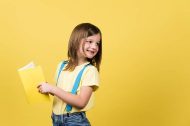 Cheerful little girl holding book and looking back at yellow copy space.
