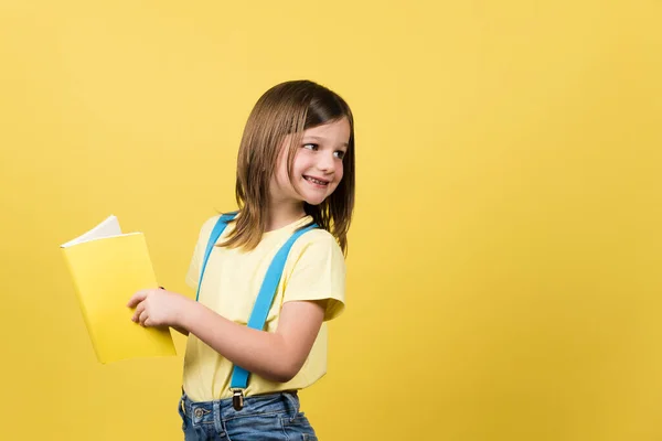 Cheerful Little Girl Holding Book Looking Back Yellow Copy Space — Stockfoto