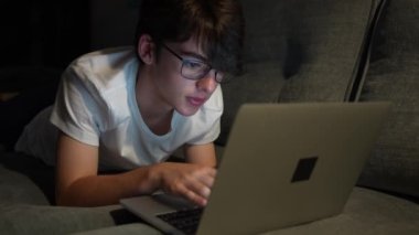 Teenager nerd boy typing on laptop. Tired cute boy working at night laying on couch. Overworking concept