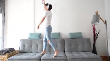 Happy teenager boy jumping and dancing on couch while listening to music on headphones