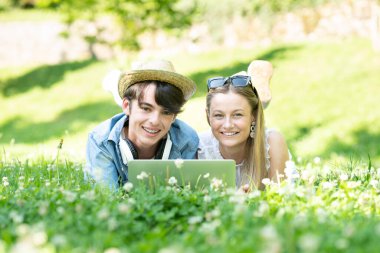 Portrait of happy teenager boy and girl lying on grass and looking at camera outdoors in a park. Students working on laptop