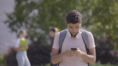 Hispanic teenager boy text messaging on phone, then taking surprise and looking at camera. Guy using social media outdoors in a park