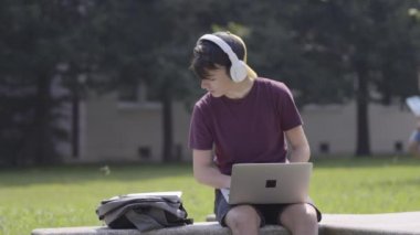 Teenager boy working on laptop outdoors while listening to music or podcast on headphones.