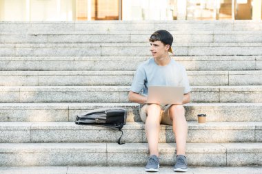 Handsome student looking at side. Teenager boy sitting on stairs working on laptop.
