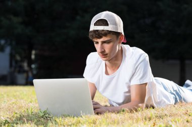 Teenager boy wearing hat working on laptop while lying down on grass, outdoors in a park.