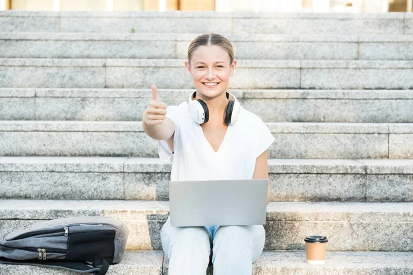 Pretty college student making ok sign. Teenager girl working on laptop while sitting on stairs