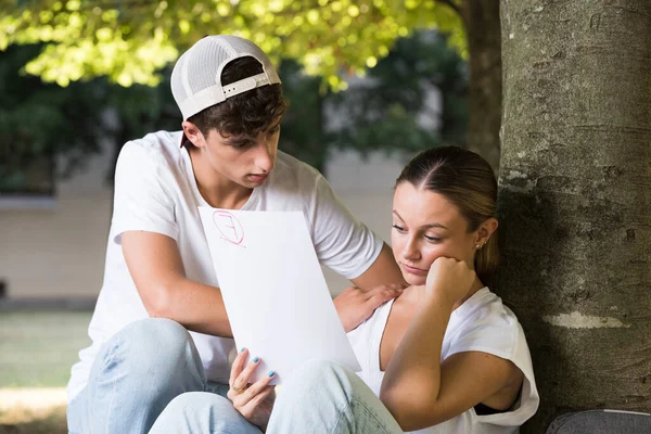 Teenager boy comforting student looking at failed exam outdoors in a park.