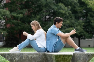Genç bir çift sırt sırta oturuyor ve parkta telefon kullanıyorlar. Dijital ve sosyal medya bağımlılığı kavramı.