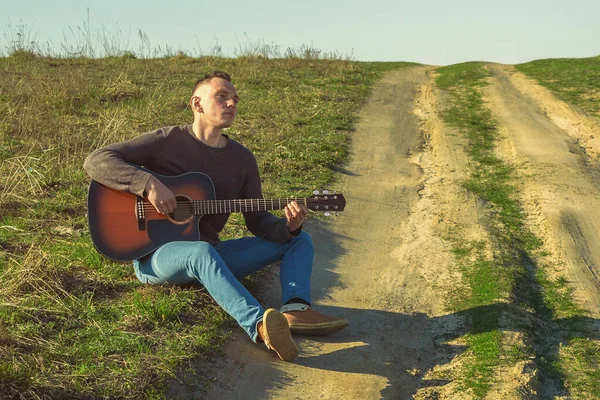 Bir adam yolda oturuyor ve akustik gitar çalıyor.