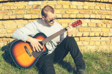 Gitarlı genç bir rockçı. Açık havada hem de. Tuğla duvarın yanında.
