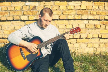 Genç bir adam gitar çalar. Gitarın yanında dinlen. Tuğla duvarın yanında.