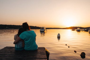 Anne ve kızı ahşap bir iskelede oturuyor, birbirlerine sarılıyor, denizin üzerindeki batan güneşe hayranlıkla bakıyorlar.