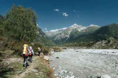 Büyük sırt çantalı bir grup turist Kafkasya Dağları 'ndaki karlı tepelere tepeden bakan çimenli yamaca tırmanıyor.