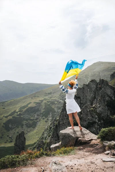 A happy woman stands near the national Ukrainian flag on the top of a mountain. View of the Carpathians on a sunny summer day.The concept of hiking, determination, self-realization, achieving goals.