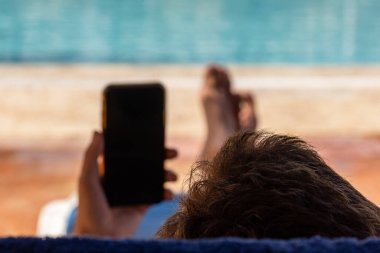 Man lies down near the swimming pool and holds a smartphone in his hand.