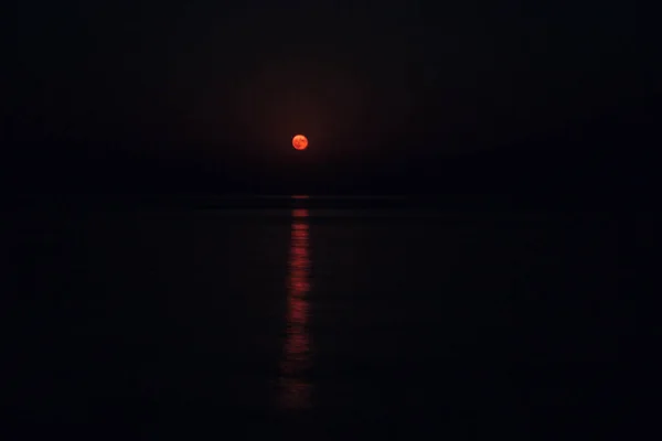 Full röd måne lyser klart i himlen på en mörk natt över det lugna havet — Stockfoto
